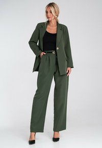 Zielona dopasowana marynarka w zestawie z szerokimi spodniami. Zawiera zapięcie na guzik, kieszenie oraz czarną bluzkę. Całość dopełniają czarne czółenka.