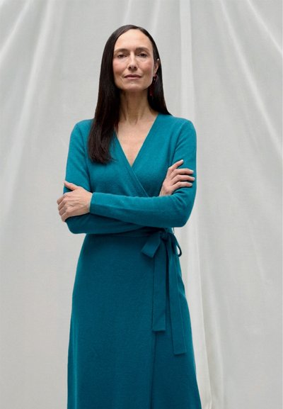Mujer de cabello largo y oscuro, de pie con los brazos cruzados, vistiendo un vestido cruzado color verde azulado sobre un fondo claro y sencillo.