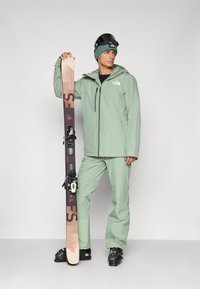 Equipo de esquí que incluye una chaqueta y pantalones impermeables de color verde claro, sosteniendo esquís negros y marrones con fijaciones, complementados con un gorro oscuro.