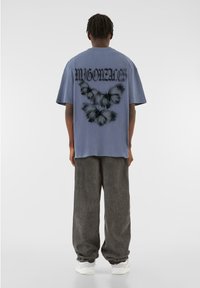 Tricou supradimensionat gri, cu un fundal albastru, grafică mare cu o fluture și textul "MYGONAE" imprimat cu litere îndrăznețe negre pe spate.