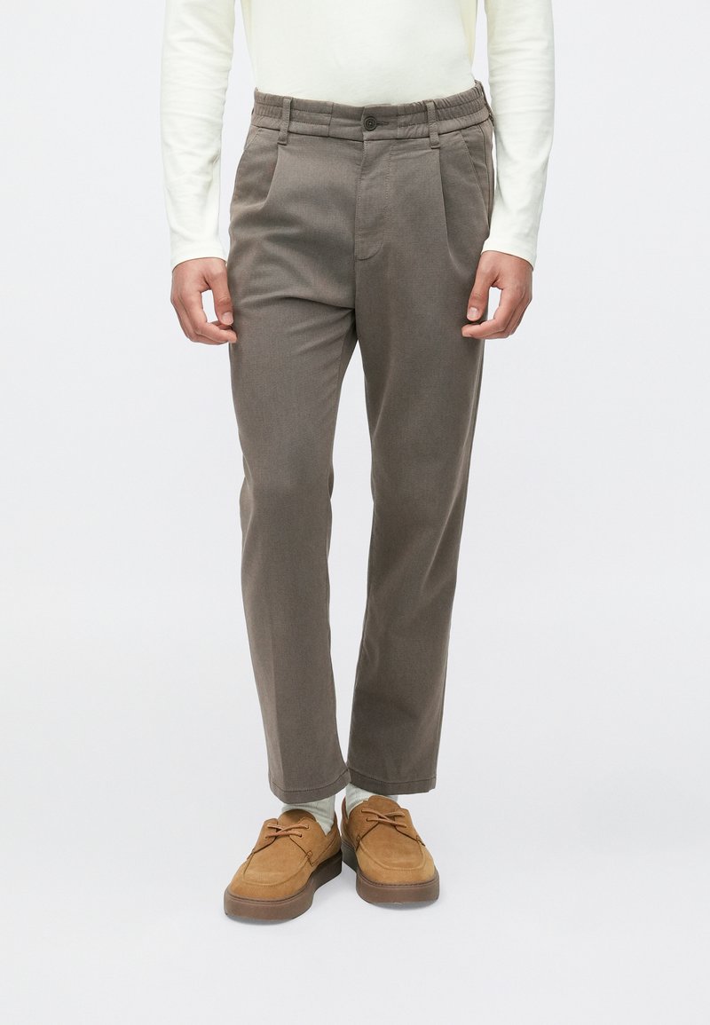 Brązowe, zwężane spodnie z fakturowanej tkaniny, z elastycznym paskiem i zapięciem na guzik. Noszone z brązowymi butami i zielonkawymi skarpetkami.