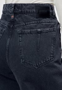 Personne portant un jean noir taille haute avec des poches arrière visibles et une étiquette en cuir noir de la marque Drykorn à la ceinture.