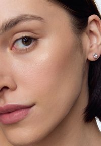 Srebrna kolczyk z przezroczystym, okrągłym kamieniem szlachetnym wyeksponowanym w centrum. Zbliżenie na ucho z gładką skórą i subtelnymi tonami makijażu.