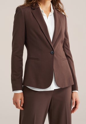 Kvinde iført en brun skræddersyet blazer med en sort knap og matchende bukser over en hvid skjorte med knapper.