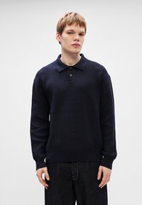 Pulôver tricotado azul marinho com colarinho tipo polo, três botões, textura canelada e mangas longas. Usado com calças de ganga escuras.