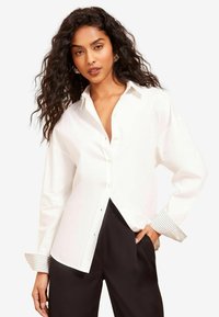 Chemise blanche à boutons avec un col, dotée de poignets rayés. Fabriquée en tissu lisse, avec une coupe décontractée et des boutons visibles sur le devant.