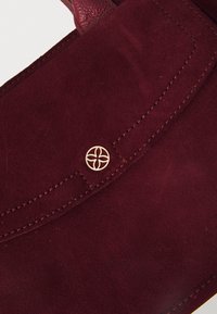 Bordeaux farvet suedetaske med en glat tekstur, foldet klap med gylden logo accent og kontrast syninger.