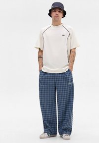 Light-colored short-sleeve shirt with black accents, paired with blue plaid wide-leg pants. Worn with casual white shoes and a bucket hat.