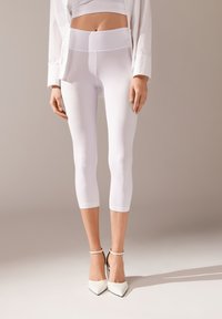Leggings blancos de cintura alta con una textura suave y longitud hasta el tobillo, combinados con tacones blancos de punta afilada, sobre un fondo neutro.