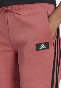 Lichtroze joggingbroek gemaakt van zachte stof, met zwarte zijstrepen en een Adidas-logo patch op het voorvak.