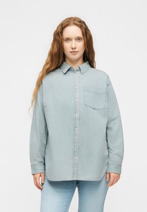 Jonge vrouw met lang rood haar, gekleed in een lichtblauw oversized spijkerblouse en bijpassende jeans, staand tegen een effen achtergrond.