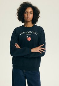 Kobieta o kręconych włosach, ubrana w czarną bluzę z napisem "POMODORO Ciliegino" i czerwonym nadrukiem pomidora, stojąca z założonymi rękami.