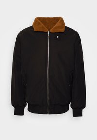 Veste noire avec un col en peluche marron. Elle possède une fermeture éclair à l'avant, des poignets côtelés et des poches latérales. Fabriquée dans un matériau lisse et durable.