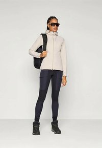 Giacca a zip beige chiaro, leggings neri con dettagli testurizzati, robuste scarpe da trekking nere e uno zaino nero. Gli occhiali da sole completano il look.