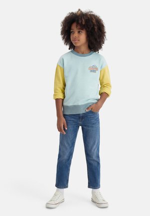 Child with curly hair wearing a blue and yellow sweatshirt, blue jeans, white socks, and white sneakers standing with one hand in pocket.