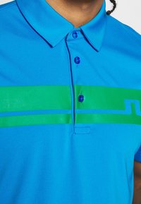 Blå polo t-shirt med ett grönt horisontellt rand och tunna blå detaljer. Tillverkad av mjuk väv med knapplist och krage.