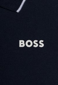 Námorníckomodrý pletený materiál s textúrovaným povrchom, na ktorom je výrazne zobrazené biele logo "BOSS". Jednoduchý dizajn goliera s kontrastnými detailmi.
