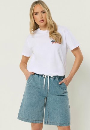 Lyshåret kvinde iført en hvid T-shirt med grafik kombineret med løstsiddende denimshorts med snøre i taljen, hænderne i lommerne.