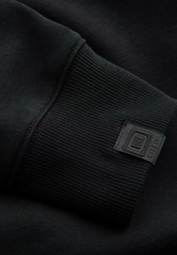 Poignet en tissu noir à texture côtelée avec une étiquette rectangulaire noire portant le mot "endle". Détail de couture visible le long du bord.