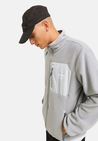 Chaqueta de forro polar gris con un alto cuello, bolsillo en el pecho con cremallera y acentos de cremallera azul. Combinada con una gorra negra. Diseño simple y funcional.