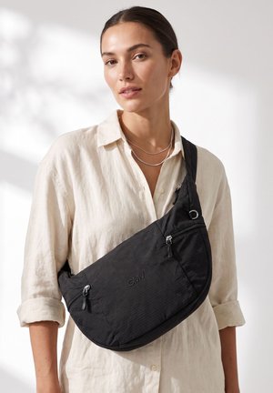 Femme portant une chemise crème boutonnée et des colliers argentés superposés, tenant un sac bandoulière noir avec le logo GMV et des poches zippées.