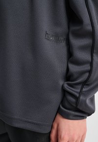 Mörkgrå idrottströja med lång ärm och textilstruktur. Har en diskret logotyp nära fållen och kontrasterande sömmar längs ärmarna.