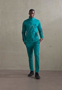 Man walking indoors wearing teal Adidas tracksuit with black stripes and black sneakers against plain beige wall.