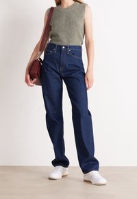 Top sin mangas de color verde oliva, combinado con vaqueros de cintura alta de color azul oscuro y zapatillas blancas. La modelo sostiene un bolso burdeos.