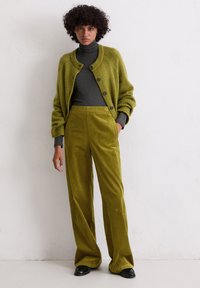 Olijfgroene gebreide vest met knopen, gecombineerd met bijpassende corduroy wijde broek. Turtleneck onderhemd in donkergrijs. Zwarte schoenen.