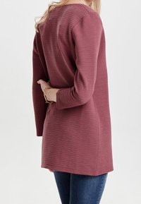 Tunika z długim rękawem w burgundowym prążkowanym materiale, z zaokrąglonym dekoltem i luźnym krojem, noszona z ciemnoniebieskimi dżinsami. Pokazano widok z tyłu.