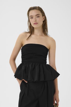 Frau mit nassen Haaren, die ein schulterfreies schwarzes Oberteil mit Schößchen an der Taille und schwarze Hosen trägt, steht vor einem einfarbigen hellen Hintergrund.