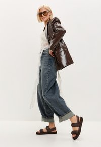 Hnědý lesklý oversized blazer přes bílý krajkový top, středně vysoké modré džíny a hnědé sandály se širokými pásky na jednobarevném pozadí.