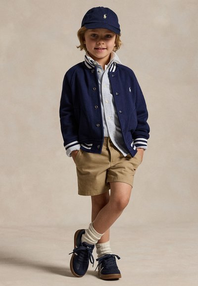 Casaco varsity de algodão azul-marinho com riscas brancas, camisa riscada azul-claro, calções caqui, sapatilhas azul-marinho e um boné azul-marinho com um emblema.