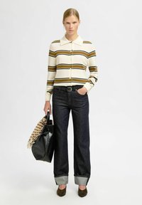 Žena u krem i smešim prugastom dugmadom džemperu, tamnim manžetama farmericama, smešim cipelama, drži crnu torbu s prugastim šalom.