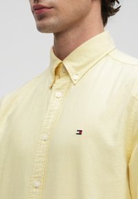 Mand iført en lysegul skjorte med knapper og lille rød, hvid og blå rektangulær logo på brystet, stående mod en ensfarvet baggrund.