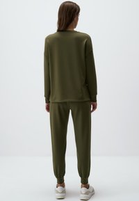 Set di abbigliamento loungewear verde oliva composto da una maglia a maniche lunghe dal fit rilassato e pantaloni a gamba affusolata con polsini a coste. Materiale morbido e elastico, design minimalista.
