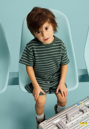 Nuori lapsi, jolla on sotkuiset ruskeat hiukset ja vihreä raidallinen asu, istuu vaaleansinisellä tuolilla lähellä hopeista matkalaukkua, jossa on tarroja.