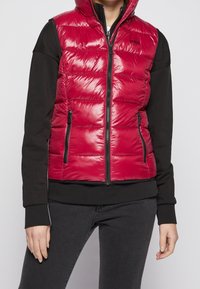 Rode puffer vest met een glanzende afwerking, rits aan de voorkant en twee zijzakken. Heeft een capuchon en een contrasterend zwart langslavend shirt eronder.