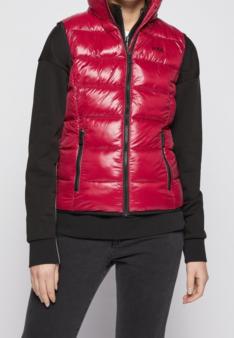 Rode puffer vest met een glanzende afwerking, rits aan de voorkant en twee zijzakken. Heeft een capuchon en een contrasterend zwart langslavend shirt eronder.