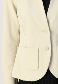 Blazer de couleur crème avec un tissu texturé, deux poches avant et deux boutons clairs. Coupe ajustée avec un design de col structuré.