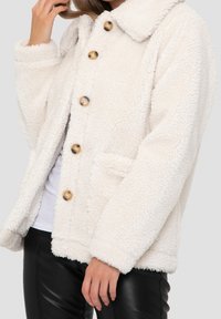 Weißes Fleece-Jackett mit texturiertem Oberflächen, übergroßem Kragen, goldenen Knöpfen und seitlichen Taschen, getragen über einem weißen Oberteil und schwarzen Hosen.