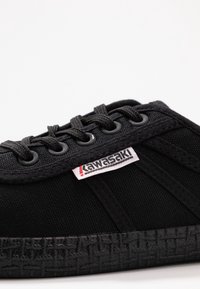 Baskets noires en toile avec un design à lacets, semelle en caoutchouc, stries texturées et étiquette logo rouge et blanche.