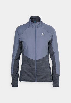 Mittelschwere Zip-Jacke in Blautönen, mit hohem Kragen und kontrastierenden, strukturierten Stoffpaneelen, seitlichen Taschen und Logodetails.