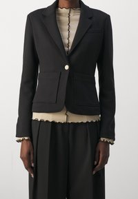 Černý saka na míru s dvěma předními kapsami, zapínání na jeden knoflík a klopovým límcem, vrstvený přes béžový žebrovaný top s vlnitými okraji.