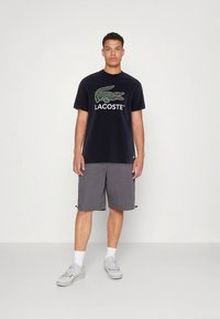 Juoda medvilninė marškinėlių su siuvinėtu žaliu krokodilo logotipu ir žodžiu „LACOSTE“. Derinama su pilkomis šortomis. Balti sportbačiai ir kojinės.