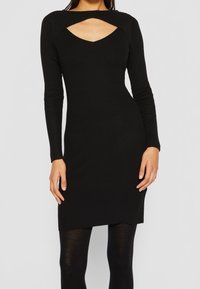 Robe noire côtelée à manches longues, dotée d'un décolleté découpé et d'une silhouette ajustée. Portée avec des collants noirs.
