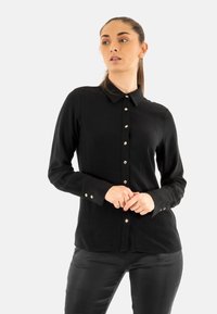 La maglietta nera a maniche lunghe presenta un collo con risvolto, una chiusura frontale con bottoni e bottoni in tonalità oro. Tessuto morbido con una texture liscia.