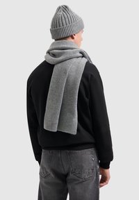 Grijze geribbelde gebreide sjaal hangt over een zwarte trui en vormt een complementair geheel met een bijpassende grijze geribbelde beanie. Model is van achteren bekeken.
