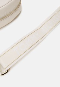 Bílá kulatá taška s texturovaným povrchem, se zlatým zipem a bílým popruhem, na kterém je v opakující se vzoru embosováno "TOMMY HILFIGER".