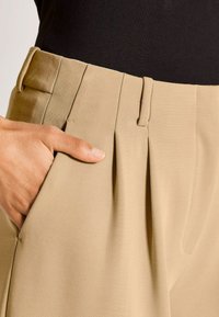 Handen rusten in de zakken van beige plooibroeken, gedragen met een zwart topje, waardoor de textuur van de stof en het op maat gemaakte ontwerp worden benadrukt.